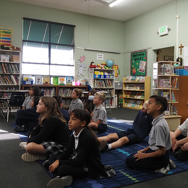 Students seated on the ground in the library, listening to the librarian read